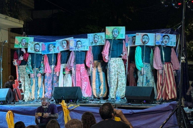 Murga performers on stage standing in a line wearing currency face cutouts that show white painted faces 